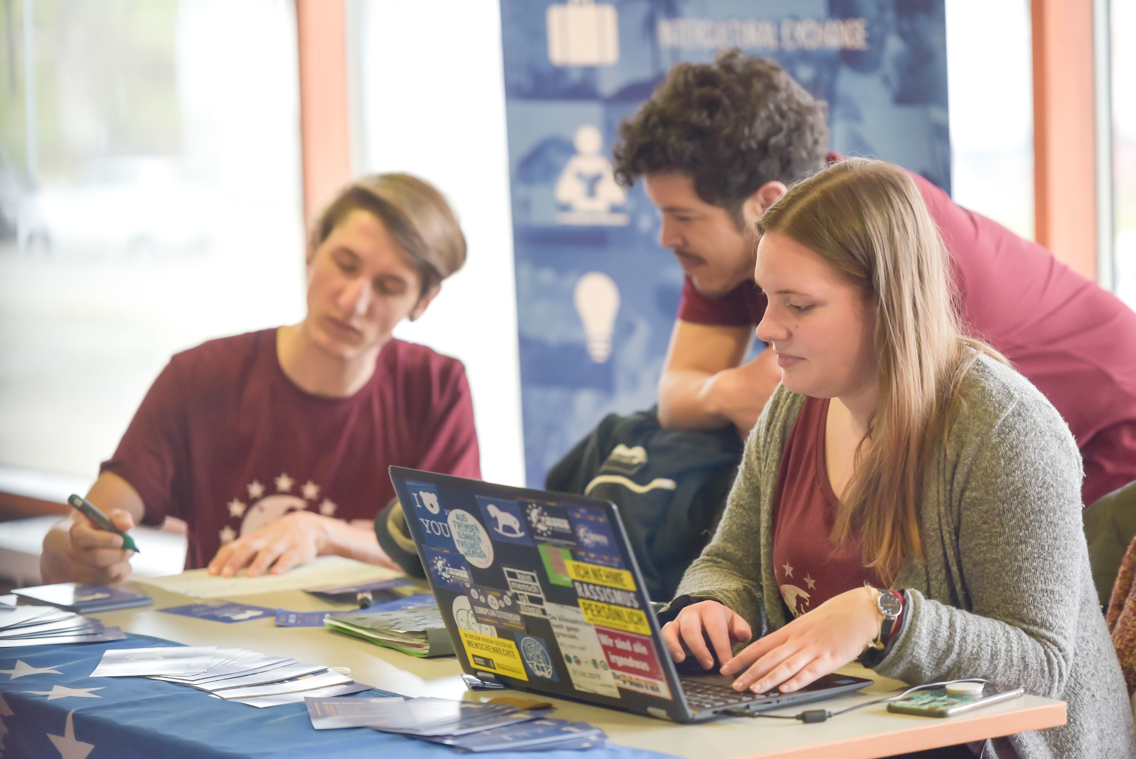 Three students are working together at an information booth. One person is sitting at a laptop, while two others are looking at documents and helping out. Information materials and flyers are laid out on the table. A roll-up banner featuring the university can be seen in the background.